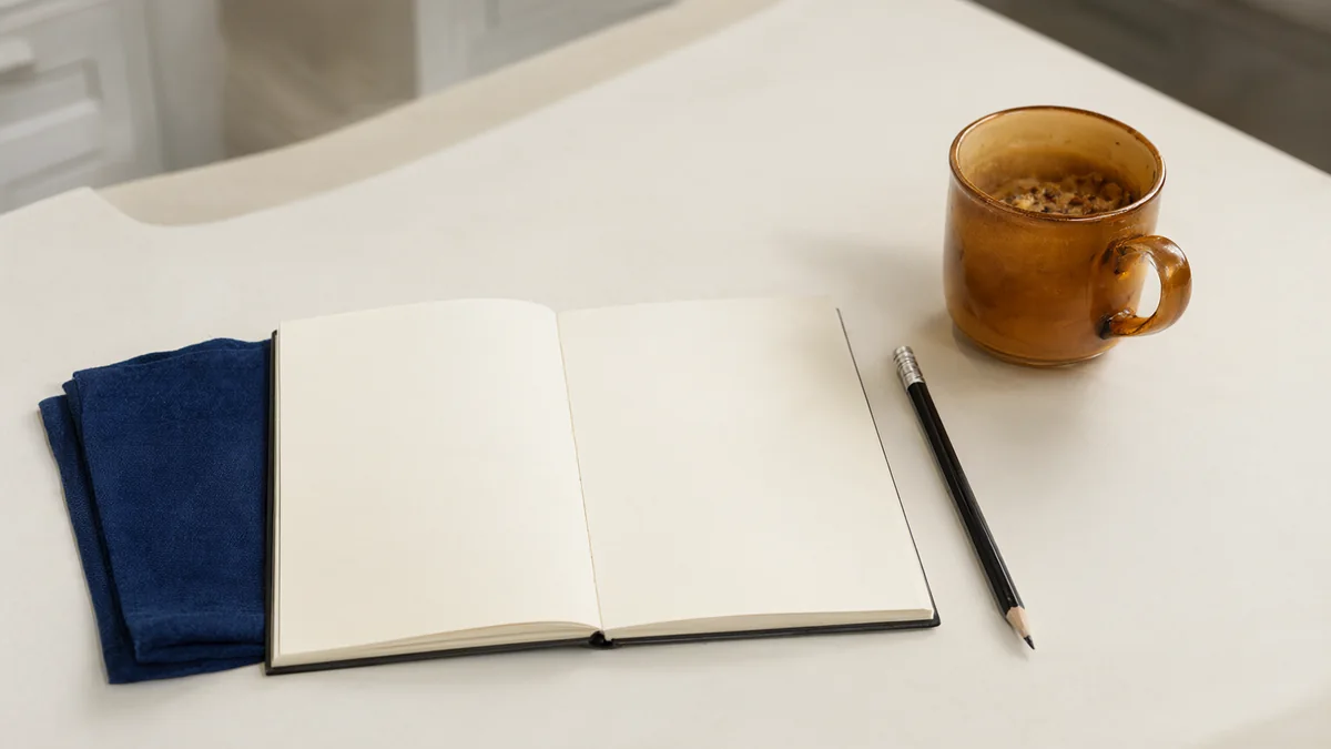 A blank journal and pencil beside a mug and folded cloth.