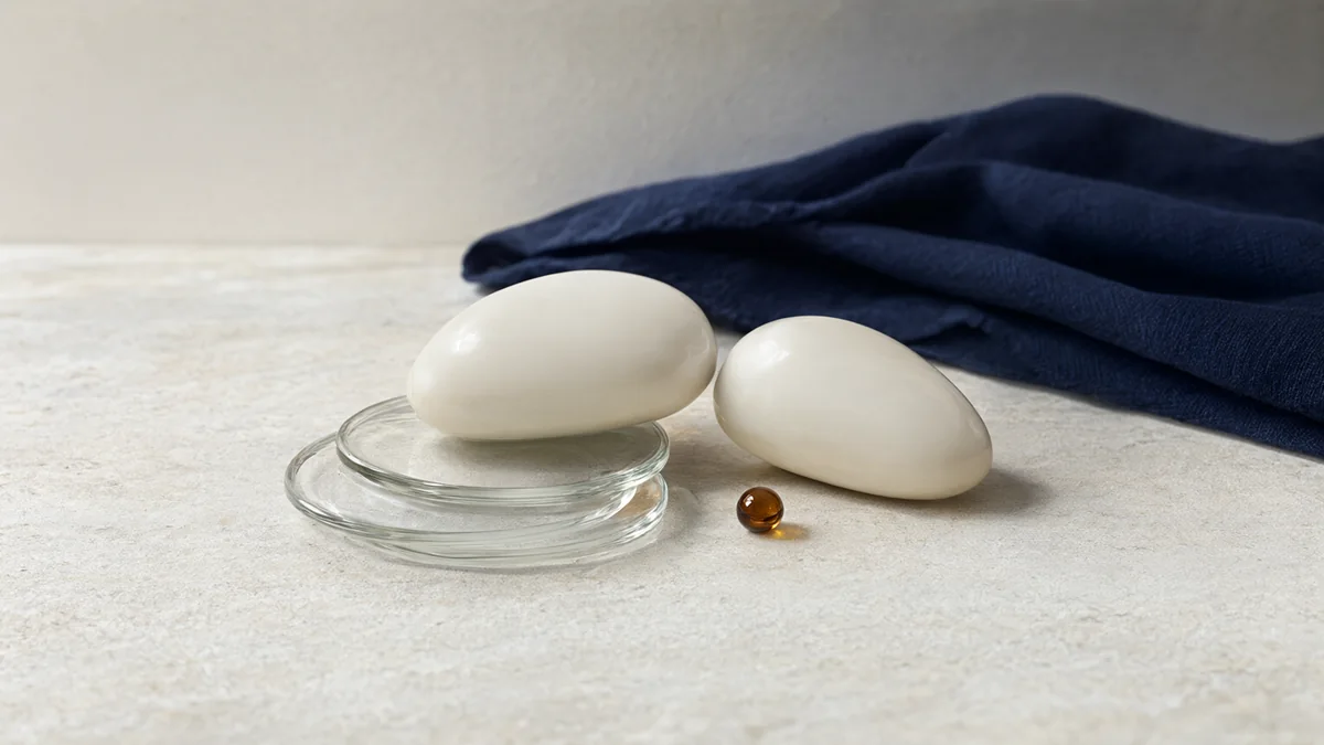 Two ivory ceramic pebbles, one on stacked glass discs, beside an amber bead and navy cloth.