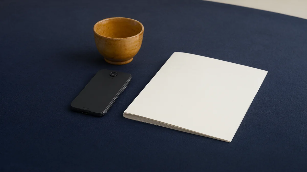 A face-down phone beside a closed notebook and ceramic cup.